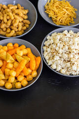 Salty snacks assortment shot on dark slate table. The picture includes potato chips, popcorn, peanut, cheese sticks, ketchup sticks, tzatziki chips, smoky flips, and others