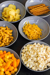 Salty snacks assortment shot on dark slate table. The picture includes potato chips, popcorn, peanut, cheese sticks, ketchup sticks, tzatziki chips, smoky flips, and others