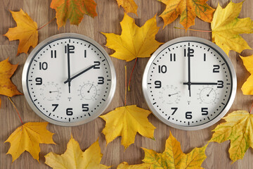 For right-to-left readers: Two clocks, one shows two o'clock, the other shows three o'clock. Yellow fallen autumn leaves lie around. Symbol of time change. Moving the hands backward from 3 to 2.