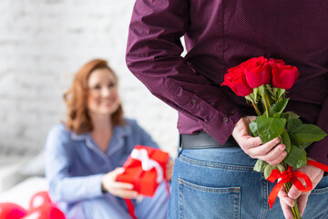 Fototapeta na wymiar Beautiful smiling young woman at home with a gift box on Saint Valentine's day. Happy day full of love, surprises. Man with red roses from the back