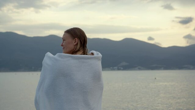 sexy slim young woman in purple swimsuit wraps herself in white towel on seashore in morning or evening. Wet girl after bathing tries to keep warm and throws towel over herself.