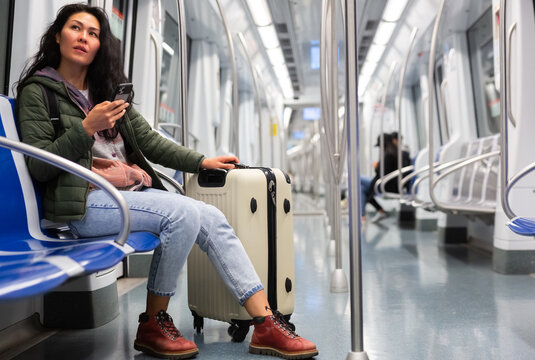 Young adult woman passenger with luggage sitting in subway car and using mobile phone