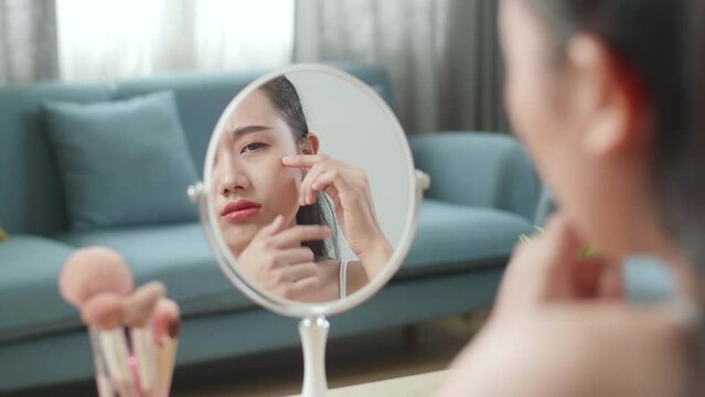 Reflection In A Mirror Worried Young Asian Woman Popping Pimples While Applying Makeup At Home
