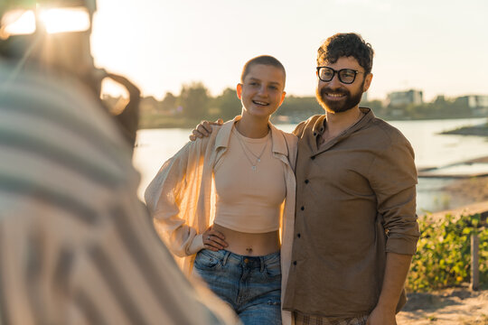 Photographer POV. Young caucasian couple asking someone to take their picture. Bald queer person in their 20s posing with caucasian bearded hipster guy by the river. Blurred unrecognizable