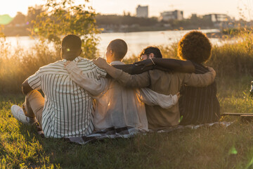 Beauty of friendship bond. Outdoor shot. Group of people sitting by the river, watching sunset, holding each others arms. Together for equality. Golden hour sunshine. High quality photo