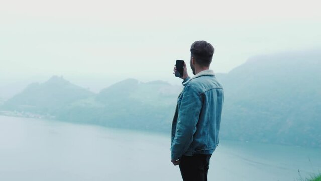 Young guy admiring swiss nature, mountains and the lake, standing and taking a photo on the cliff of a mountain. Happy excited man looking around at amazing scenery, foggy nature in autumn 