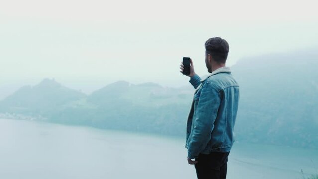 Handsome stylish guy man holding phone and recording a video or taking a photo on his modern smartphone, standing on the cliff of a mountain in Switzerland, looking at amazing foggy scenery nature 