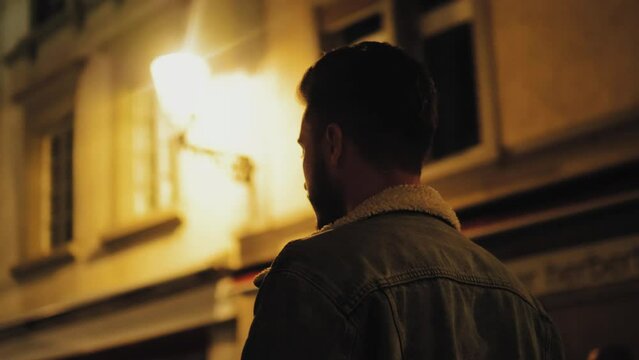 Attractive man standing in front of light, flashlight at night or in the evening, on the street. Urban atmosphere. Dark street. Back view or side view of handsome young guy student 