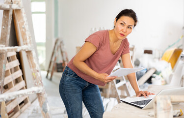 Asian woman designer checking documents and using her laptop during repair works indoors.