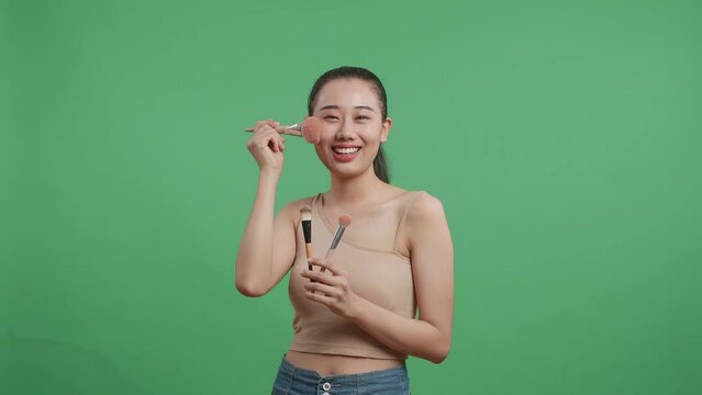 Beautiful Asian Woman Holding Makeup Brushes And Rouging Her Cheeks While Smiling To Camera On Green Screen Background In The Studio
