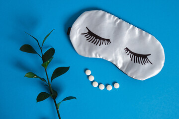 sleep mask with painted eyelashes and insomnia pills in the shape of a smile on a blue background