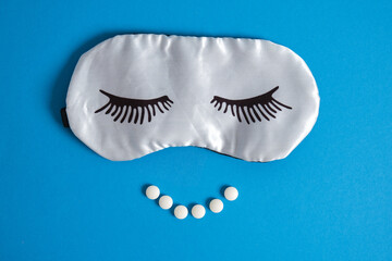 sleep mask with painted eyelashes and insomnia pills in the shape of a smile on a blue background
