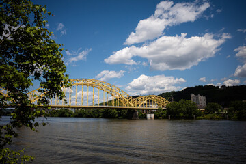 Yellow bridge crossing river in Pittsburgh with blue sky and white clouds.