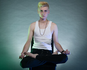 Non-Binary citizen with short cropped blonde hair, wearing black jeans and A-shirt poses in studio under multi-colored lights.