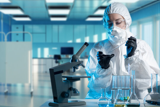 Virologist is working. Woman virologist in chemical protection suit. Girl is studying dangerous viruses. Biologist at laboratory table. Virological clinic employee. Virologist works with microscope