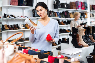 Asian woman selecting new shoews while standing in salesroom shoeshop. Elderly European woman shopping in background.