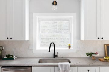 A beautiful kitchen sink detail with white cabinets, marble countertops, a hexagon tiled backsplash, and a gold and black light above the sink.