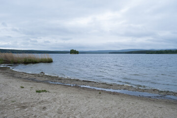 View of the sandy beach and lake on a cloudy day. Spring river landscape. Boundless beautiful sky, a blue lake with a small green island and a sandy shore. Deserted, quiet, silence. Russia, South Ural