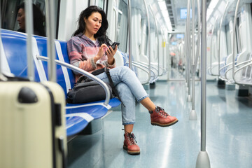 Asian woman with suitcase and sitting on bench in subway train and using smartphone