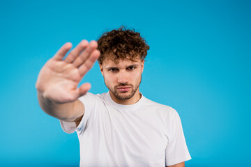 Portrait of young curly man in white t-shirt holds his hand showing stop sign on blue background