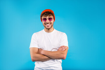 Attractive young smiling man in white t-shirt and orange hat wears glasses stands on blue background