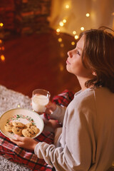 Caucasian fair hair boy waiting for Santa holding in hands glass of milk, cookies sitting on floor at home in Christmas Eve. Happy holidays time Winter spirit Getting cozy vibes Christmas decoration