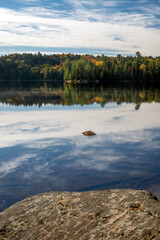 A rocky shoreline leads to a calm lake reflecting clouds and the tree lined shoreline across the water. Harness Lake, Algonquin Provincial Park, Ontario, Canada.