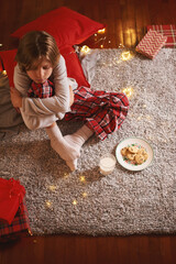 Cute tired child waiting for Santa with glass of milk and cookies sitting on floor at home in Christmas Eve. Christmas decoration. Candid moment Winter time season Dreamy look about miracle Top view