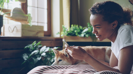 Pretty girl student is using smartphone touching screen and laughing lying on bed at home with cute pet dog, animal is enjoying care and love. Technology and animals concept.