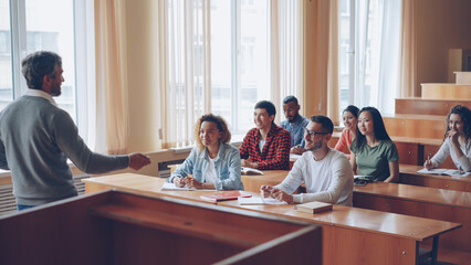 Experienced high school teacher is talking to cheerful students while attractive young people are listening, smiling and laughing sitting at desks in light classroom.