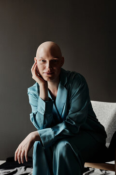 Portrait of elegant bald woman in silk suit sitting on armchair and looking at camera