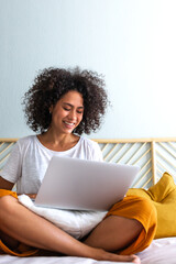 Fototapeta na wymiar Multiracial latina woman using laptop sitting on bed at home. Vertical image. Copy space. Technology concept.