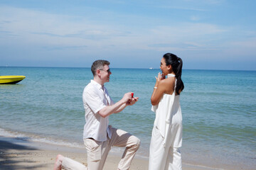 Surprised man kneeling holding a ring to propose to his girlfriend on the beach bright blue sea.