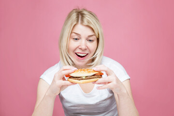 A young woman happily eats a hamburger. Beautiful blonde in a white shirt. Hunger, diets and junk delicious food. Pink background.