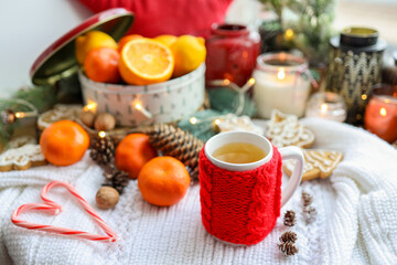 A cup of tea with a knitted red decoration, tangerines, candles, a garland. Christmas atmosphere in the house