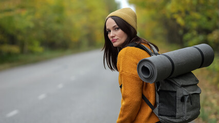 Adult woman with backpack stands near empty road and waits for bus to start journey. Hiker wearing hat waits for vehicle in thick autumn forest