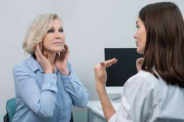 Young female cosmetologist gives advice on anti-ageing facial injections. Dark-haired doctor touches blonde woman wrinkles and sketches notes in patient form