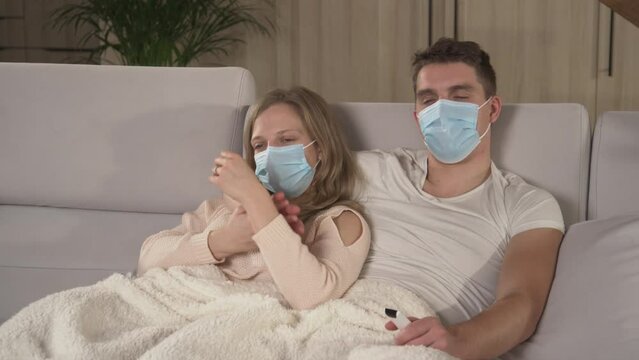 CLOSE UP: Couple wearing protective face masks and watching TV during sick leave. Autumn colds and flu spreading around. Young couple getting over seasonal flu while lying on cosy couch under blanket.