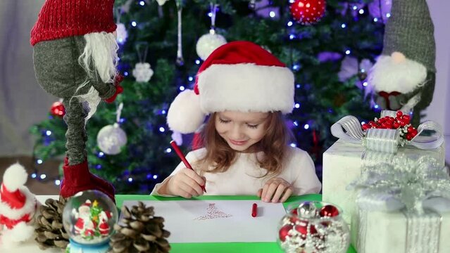 A little girl in a red Santa's Hat writes a letter to Santa Claus with her wishes under the Christmas tree. Christmas and New Year atmosphere