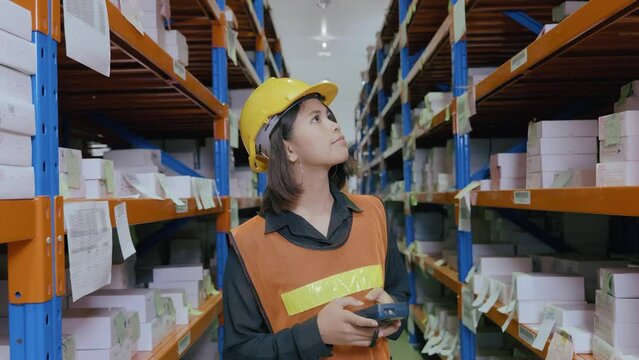 A female worker in uniform with her safety helmet on is walking by holding scanner for scanning parcel barcode in the warehouse store background. Inventory digitalization with barcode scanner concept.