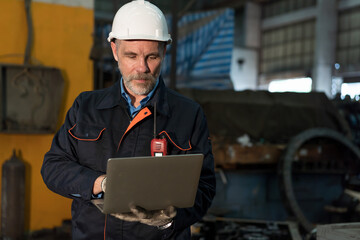 Senior male engineer working with laptop computer at the industry factory. Male technician wear safety helmet, gloves and uniform working in the factory
