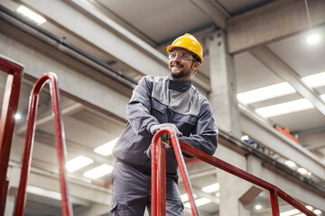 A smiling factory supervisor in a protective uniform stands on the height and monitors work in a...