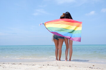 A lesbian couple holding a rainbow flag LGBT symbol on the beach against a bright blue sky background. LGBT Concepts.