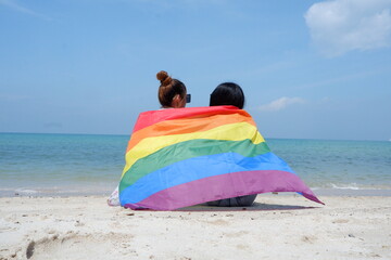 A lesbian couple holding a rainbow flag of LGBT symbols sits on the beach against a bright blue sky background. LGBT Concepts.