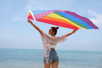 Young woman holding a rainbow flag LGBT symbol on the beach against a bright blue sky background. LGBT Concepts.