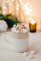 Christmas mug with hot chocolate and marshmallow with cozy garland lights and christmas tree branches on the background