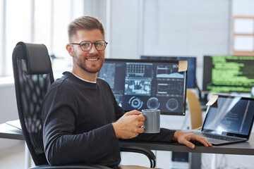 Portrait of software programmer smiling at camera while posing at workplace in office with coffee...