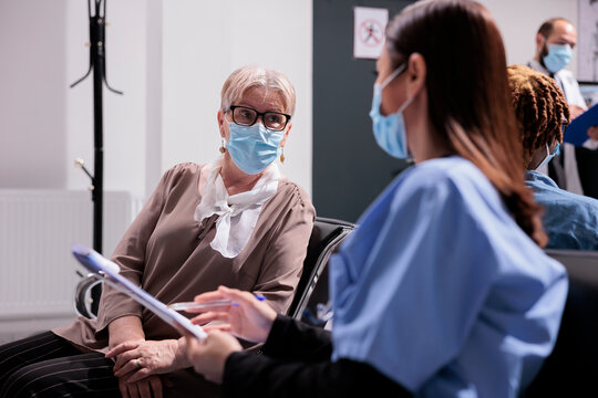 Elderly Woman Being Treated By Asian Medical Practitioner In Sanatorium. Medium Shot Of Patient At Busy Clinic, Medical Consultation. Female Doctor Interviewing Senior Female Patient At Waiting Room.