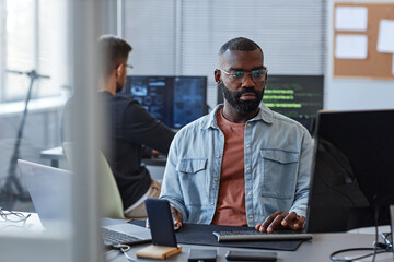 Portrait of black man using computer while programming mobile software in office behind glass wall
