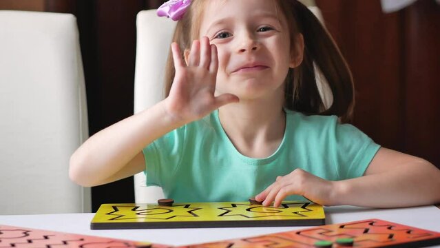 A little girl at a table with an interhemispheric board looks into the frame smiles and waves her hand affably. Correctional classes for children with mental retardation and hyperactivity.